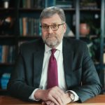 Portrait of BOV member Chris Tuttle sat at a desk in front of a bookshelf.